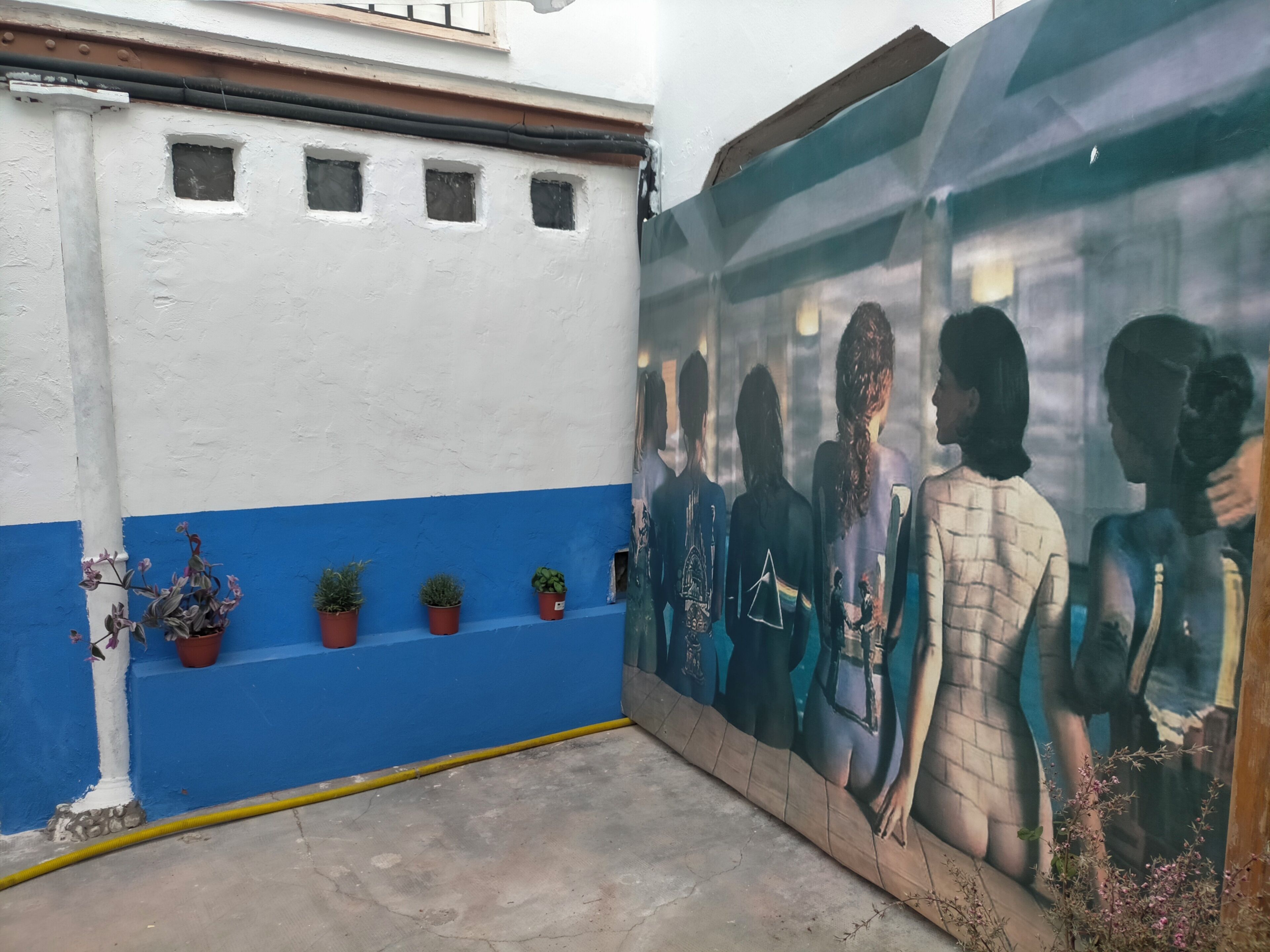 Un edificio con fachada azul y blanca, plantas en macetas en la pared baja y un mural a la derecha.