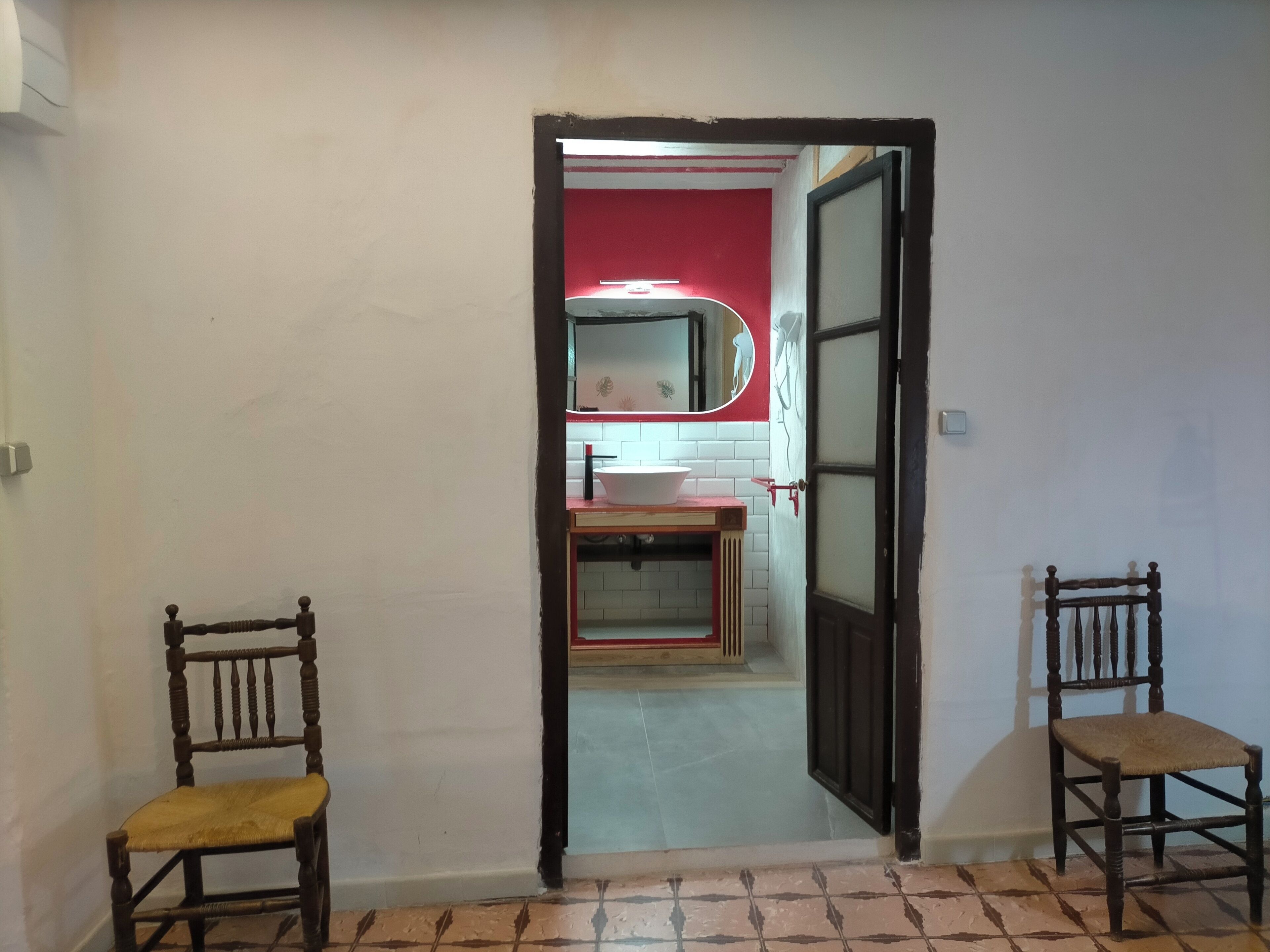 Een gang die leidt naar een badkamer met een rode muur, een wit wastafel en een spiegel. In de gang staan twee houten stoelen.