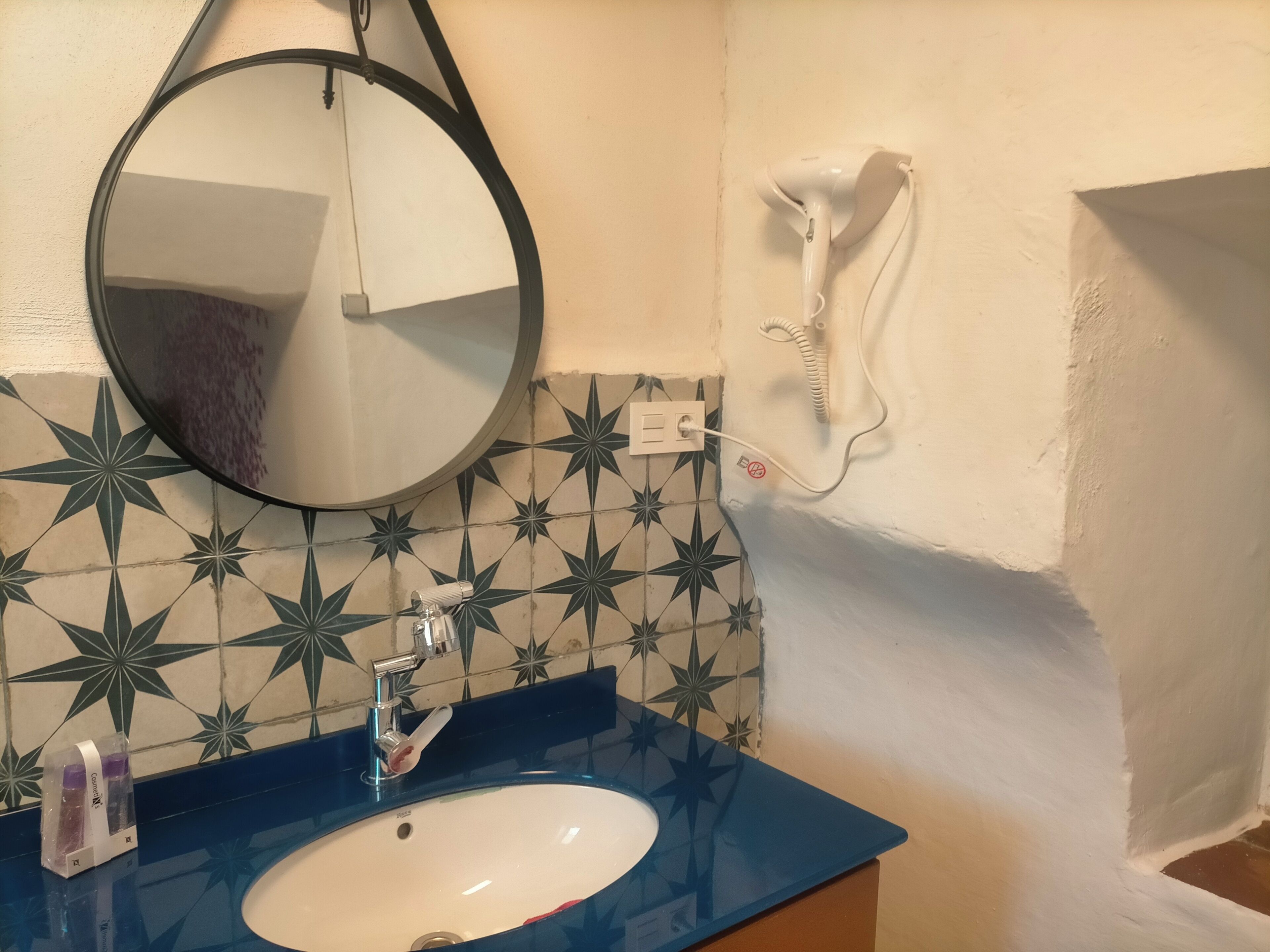 Een badkamer met een ronde spiegel, een wastafel, een haardroger en een betegelde achterwand.