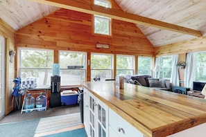 A cabin interior with a wooden ceiling, a kitchenette, a seating area with a sofa, and large windows.