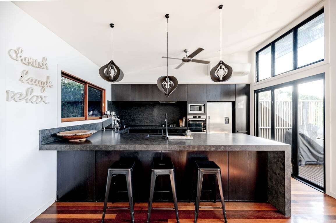 A modern kitchen with a central island, bar stools, pendant lights, a ceiling fan, and a large window.