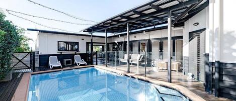 A modern outdoor pool area with clear glass walls, lounge chairs, and a covered seating area.
