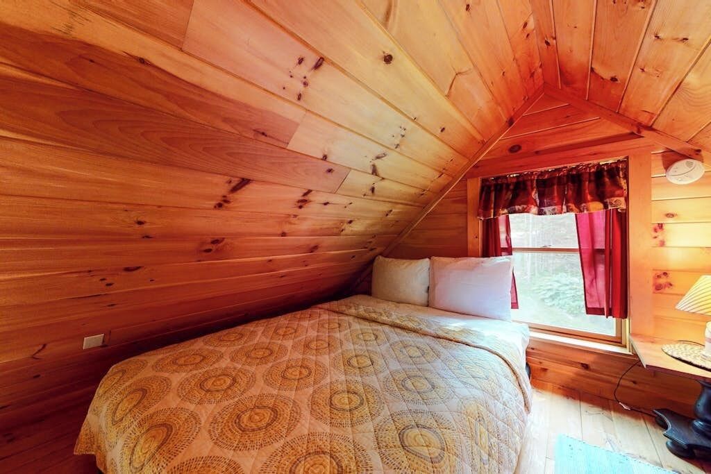 A cozy room with wooden walls, a bed with patterned bedding, a window with curtains, and a lamp on a bedside table.