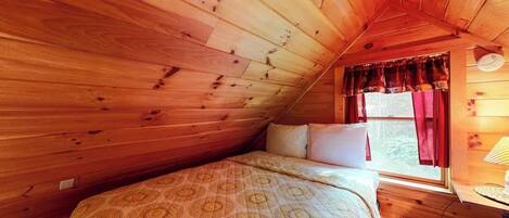 A cozy room with wooden walls, a bed with patterned bedding, a window with curtains, and a lamp on a bedside table.