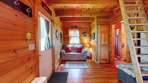 A log cabin interior with a wooden floor, a sofa, a ladder, and a door leading to another room.
