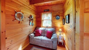 A cozy cabin interior with a wooden floor, a sofa with cushions, a small table with a lamp, a window with curtains, and a wall-mounted clock.