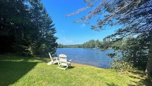 A grassy area with two Adirondack chairs facing a lake, surrounded by trees.