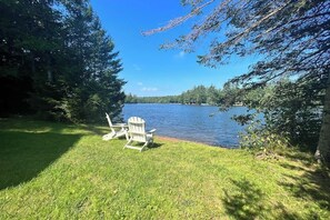 A grassy area with two Adirondack chairs facing a lake, surrounded by trees.