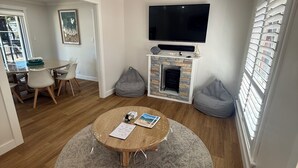 Ett vardagsrum med ett runt soffbord, en platt-TV som är monterad ovanför en stenbeklädd öppen spis och en matplats med en glasdörr som leder ut.