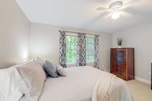 Ein Schlafzimmer mit Bett, Deckenventilator, Fenster mit gemusterten Vorhängen und einem Holzkasten.