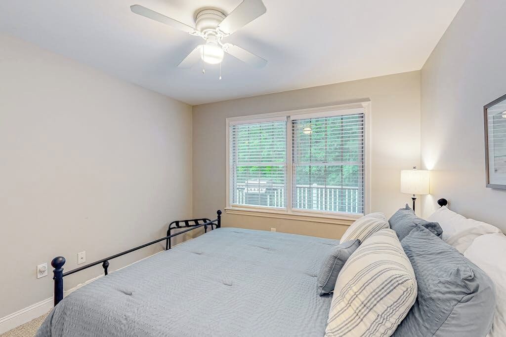 Une chambre à coucher comprenant un lit, un ventilateur de plafond, une fenêtre avec des stores et une table de chevet avec une lampe.