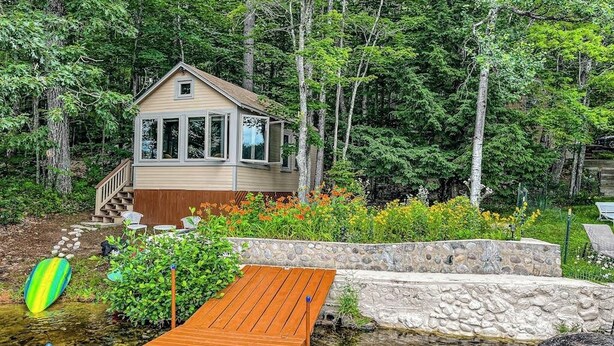 A cabin with a stone retaining wall, wooden deck, and a small garden with colorful flowers. - Refurbished waterfront 1950s cabin with sauna, firepit, grill & kayaks - dogs ok (Raymond)