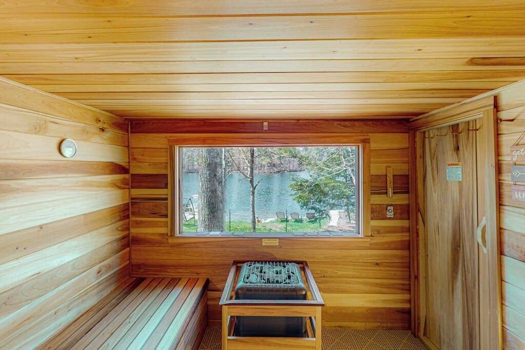 Un sauna en bois, avec un banc, une fenêtre donnant sur des arbres et un plan d’eau, ainsi qu’un appareil de chauffage pour sauna.