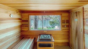 A wooden sauna room with a bench, a window with a view of trees and a body of water, and a sauna heater. - Refurbished waterfront 1950s cabin with sauna, firepit, grill & kayaks - dogs ok (Raymond)