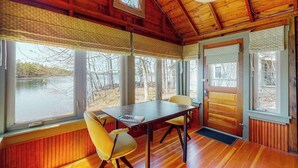 A wooden cabin with a dining table and chairs, large windows, and a view of a lake. - Refurbished waterfront 1950s cabin with sauna, firepit, grill & kayaks - dogs ok (Raymond)