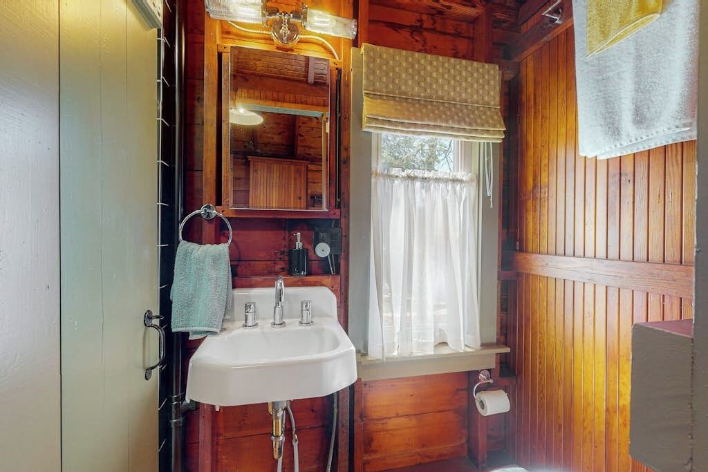 Ein Badezimmer einer Holzhütte mit Waschbecken, Spiegel und Fenster mit Vorhängen.
