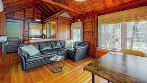 A cabin interior with a wooden ceiling, a black leather sofa, a dining table, and a kitchen area. - Refurbished waterfront 1950s cabin with sauna, firepit, grill & kayaks - dogs ok (Raymond)