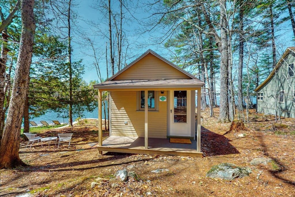 Une petite cabane dotée d’un porche, entourée d’arbres et bordée par un lac.