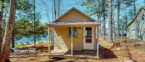 Une petite cabane dotée d’un porche, entourée d’arbres et bordée par un lac.