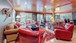 A spacious living room with a high ceiling, large windows, and a mix of seating including a red leather sofa and a white couch. - Log cabin 1 mile from Sunday River with hot tub, fireplace & Ping-Pong (Newry)