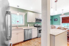 A kitchen with white cabinets, stainless steel appliances, a dishwasher, and a dining area with a table and chairs. - Sheridans' Getaway | Dog-friendly house in Newry, 1 mile to Sunday River (Newry)