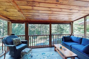 A covered outdoor deck with wicker furniture, a wooden table, and a view of the surrounding forest. - Charming Lakeview House with Deck, Cozy Fireplace and firepit (Rome)