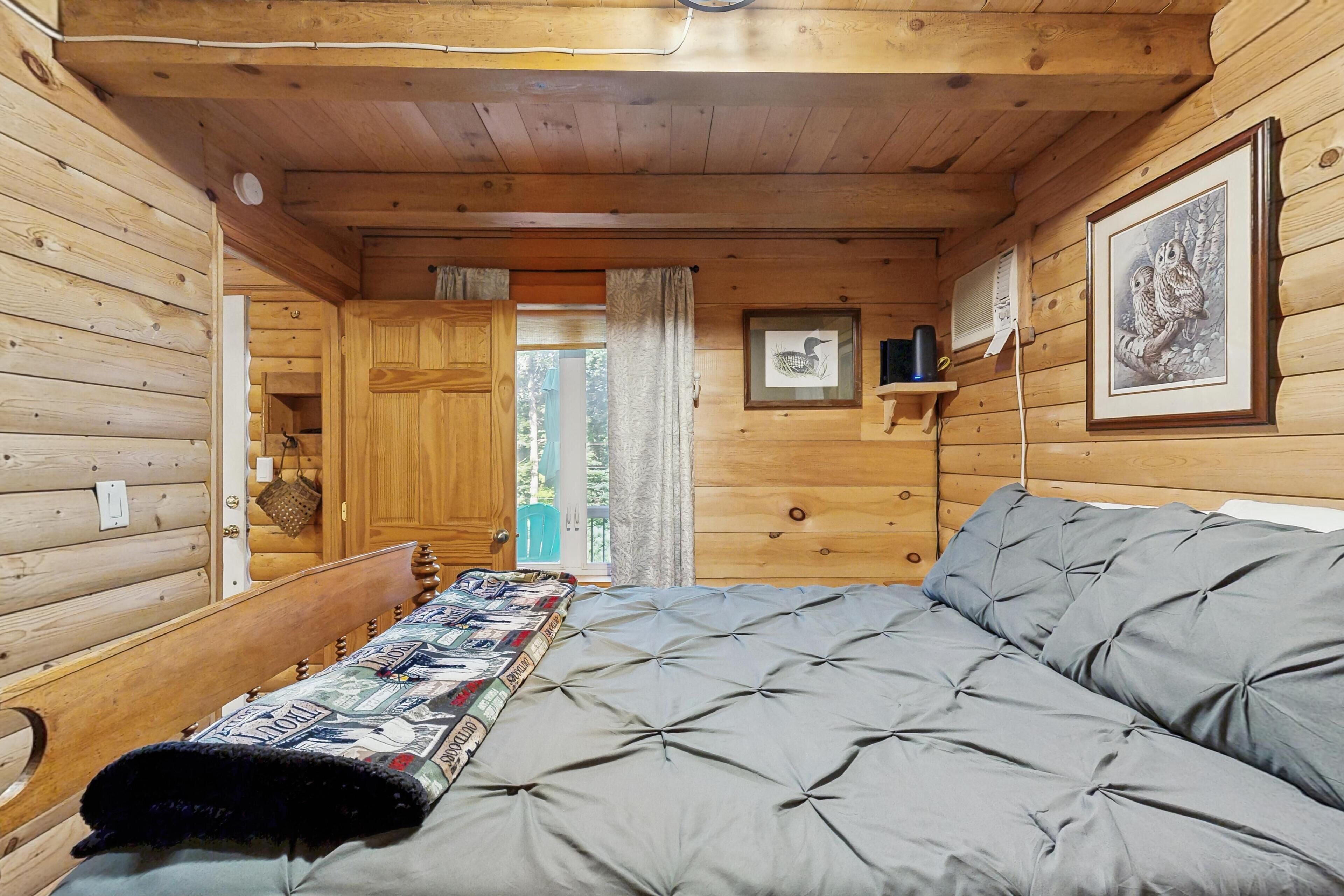 A wooden cabin interior with a bed, a door, a window, and a shelf with framed pictures.