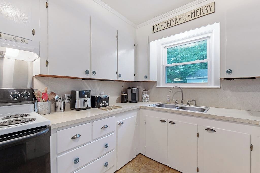 A compact kitchen with white cabinets, a stove, a coffee maker, and a window with a view of trees.