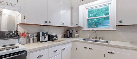 A compact kitchen with white cabinets, a stove, a coffee maker, and a window with a view of trees.