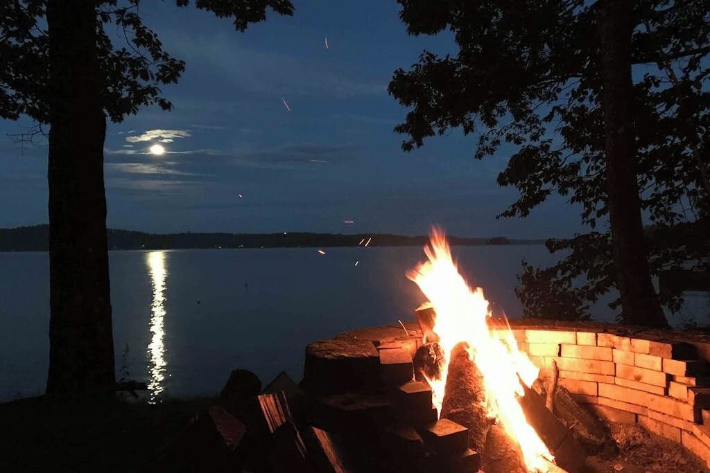 A campfire at dusk with a lake and trees in the background.