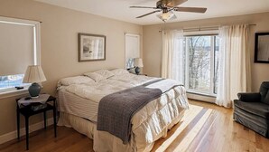 Ein Schlafzimmer mit Bett, Deckenventilator, Fenster mit Vorhängen, Nachttisch und Stuhl.