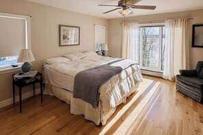 Ein Schlafzimmer mit Bett, Deckenventilator, Fenster mit Vorhängen, Nachttisch und Stuhl.