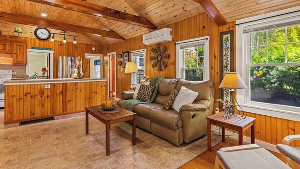 A wooden interior with a sofa, coffee table, and lamp, a kitchen area with a refrigerator, and a window with a view of greenery.