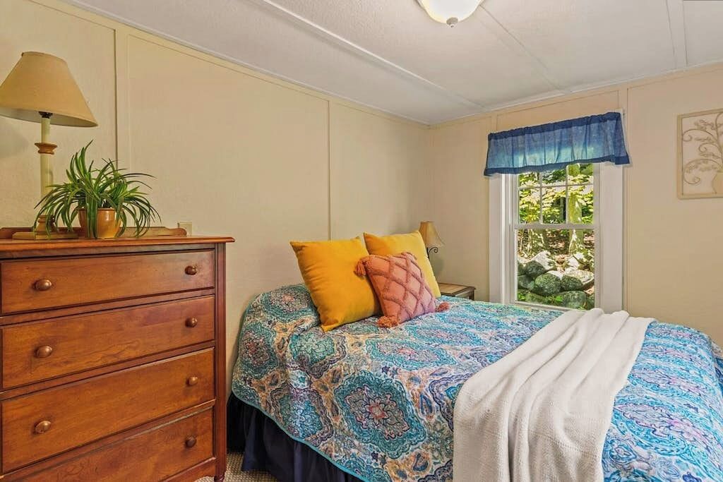 A bedroom with a bed, dresser, lamp, potted plant, window with view, and decorative wall piece.