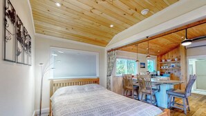 A bedroom with a bed, a desk, and a chair, wooden ceiling, and a window.