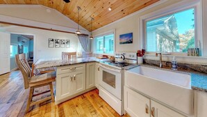A kitchen with wooden ceiling, white cabinets, a large window, and a dining area with chairs.