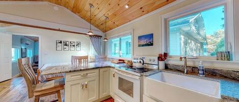 A kitchen with wooden ceiling, white cabinets, a large window, and a dining area with chairs.