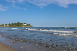 Ein Strand mit Wellen, eine entfernte Insel mit Gebäuden und ein klarer Himmel.