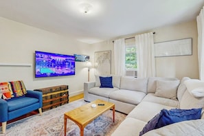 A living room with a sofa, a blue armchair, a wooden coffee table, a television, and a window with curtains. - Charming  Retreat w/ WiFi, Ping Pong Table & Walking Distance to the Ocean (Cumberland)