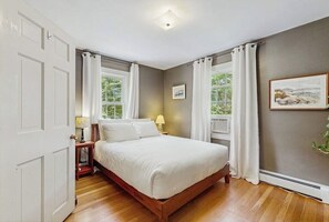 A bedroom with a bed, two bedside tables, a window with curtains, and a picture on the wall. - Charming  Retreat w/ WiFi, Ping Pong Table & Walking Distance to the Ocean (Cumberland)