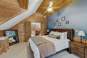 Ein Schlafzimmer mit schrägverdeckter Decke, Holzwänden, einem Bett mit weißen Leinen, Nachttischlampen und gerahmten Bildern an der Wand.
