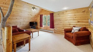 Interno di una baita con pavimento in legno, televisione, camino e un'area salotto accogliente.
