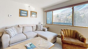 A living room with a sofa, armchair, coffee table, and large windows with a view of a mountainous landscape. - Cozy Studio with Slopeside Access, Pool, Sauna, and Mountain Views (Newry)