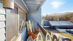 A balcony with skis leaning against the wall, a view of a snowy landscape, and a building in the background. - Cozy Studio with Slopeside Access, Pool, Sauna, and Mountain Views (Newry)