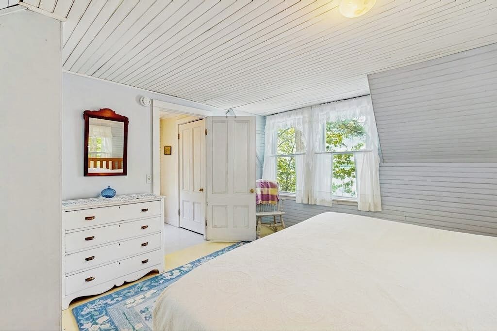 Une chambre à coucher avec un lit, une commode, une chaise et une fenêtre avec des rideaux.