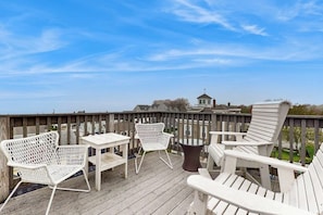 Eine Holzterrasse mit weißen Gartenmöbeln, darunter Stühle und ein Tisch, mit Blick auf eine Wohngegend.