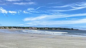 En strand med klar himmel, vågor och en avlägsen kustlinje med byggnader.