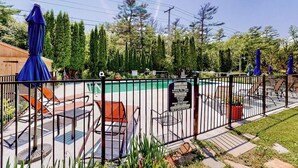 Zona de piscina al aire libre con vallas, sillas de descanso y sombrillas, rodeada de árboles.