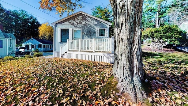 Una piccola casa a un piano con un portico, circondata da alberi e da un prato ricoperto di foglie cadute.