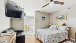 A bedroom with a bed, a desk with a chair, a television mounted on the wall, a ceiling fan, and a window with a view. - Studio condo with furnished patio, sauna, & pool - near beaches (Wells)